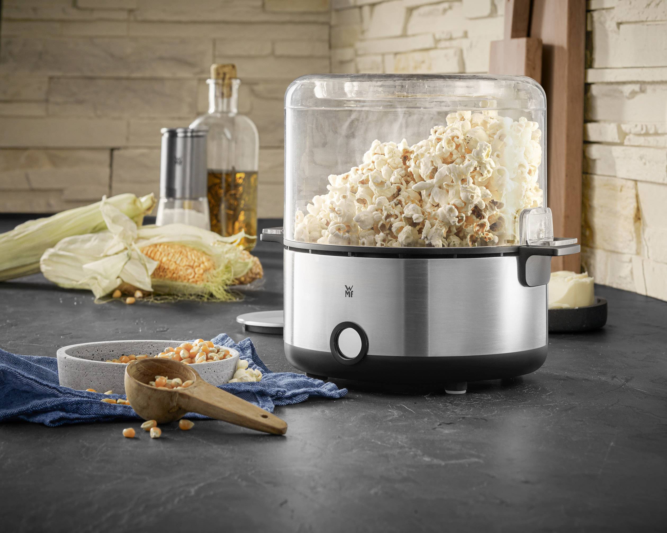 Popcornmaschine auf einer Küchenarbeitsplatte. Frisches Popcorn quillt aus der Maschine. Im Hintergrund Maiskolben und Gewürze.