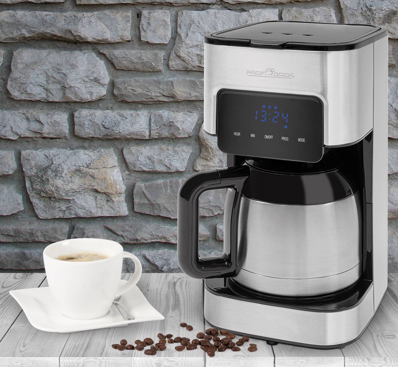 Eine silberne Kaffeemaschine auf einem Tisch, daneben eine Tasse Kaffee und verstreute Kaffeebohnen. Hintergrund aus grauem Stein.