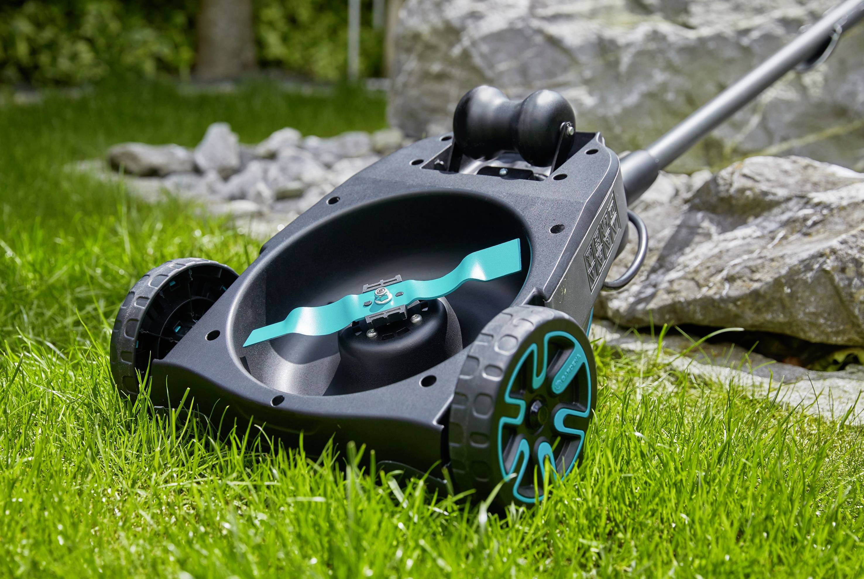 Ein Rasenmäher mit blau-schwarzem Design steht auf einer grünen Wiese. Im Hintergrund sind Steine zu sehen.