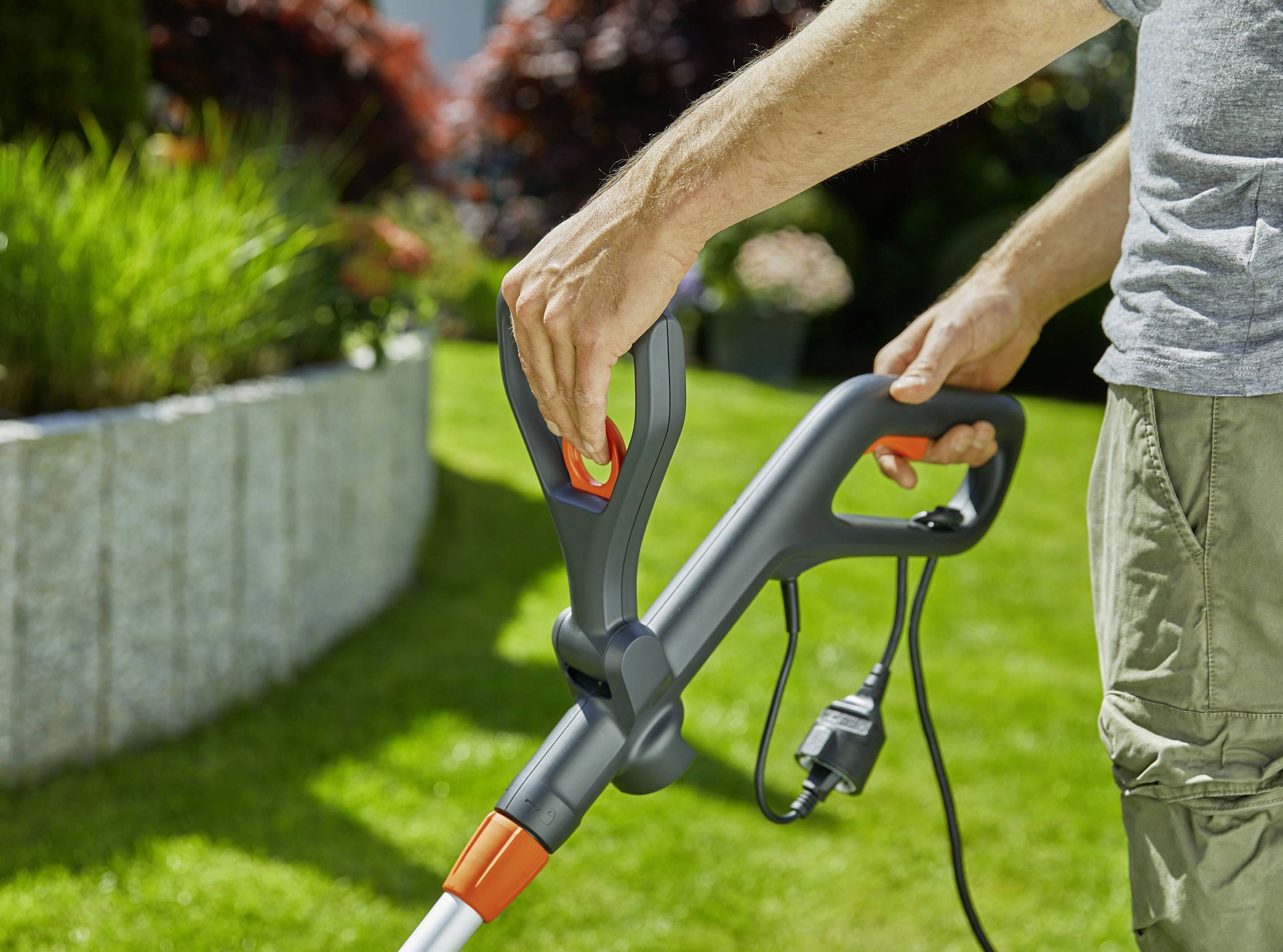 Eine Person hält einen elektrischen Rasenmäher-Griff und drückt den Startknopf. Im Hintergrund ist ein gepflegter Garten zu sehen.