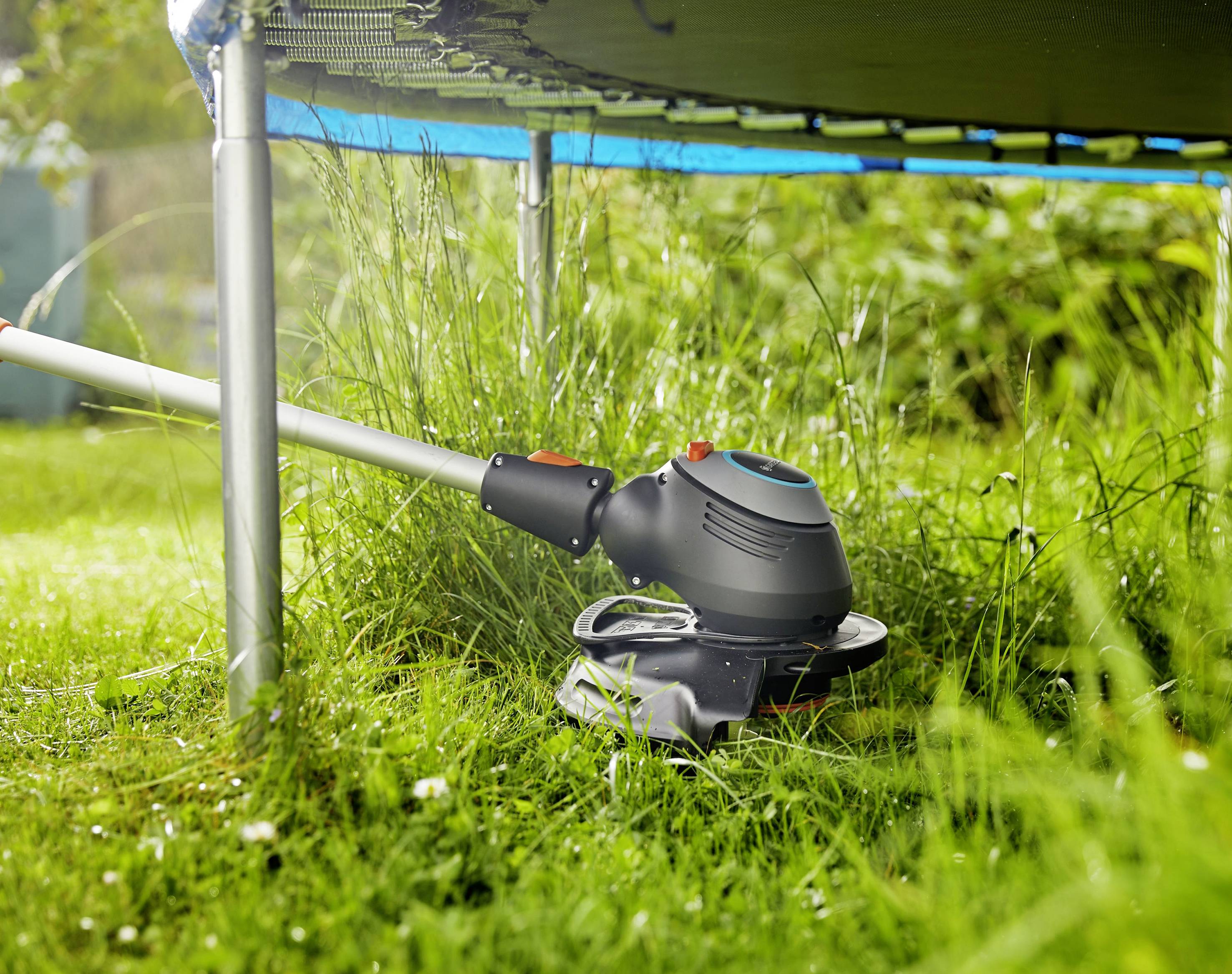 Eine elektrische Motorsense schneidet Gras neben einem Trampolin auf einem grünen Rasen. Sonnenlicht fällt auf die Szene.