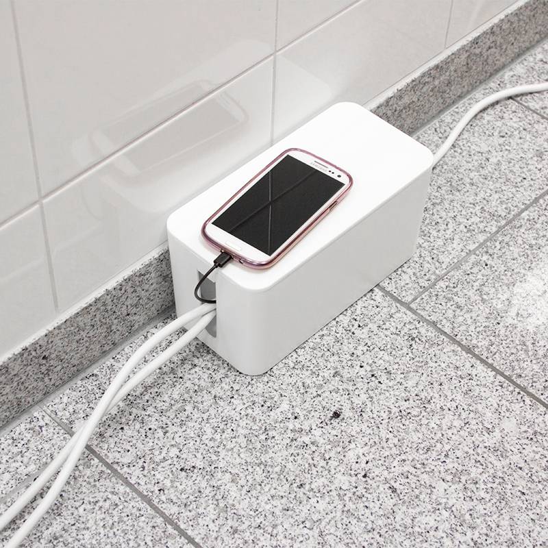 Ein Smartphone liegt auf einer weißen Kabelbox am Boden. Mehrere Kabel sind zur Box geführt, um Unordnung zu vermeiden.