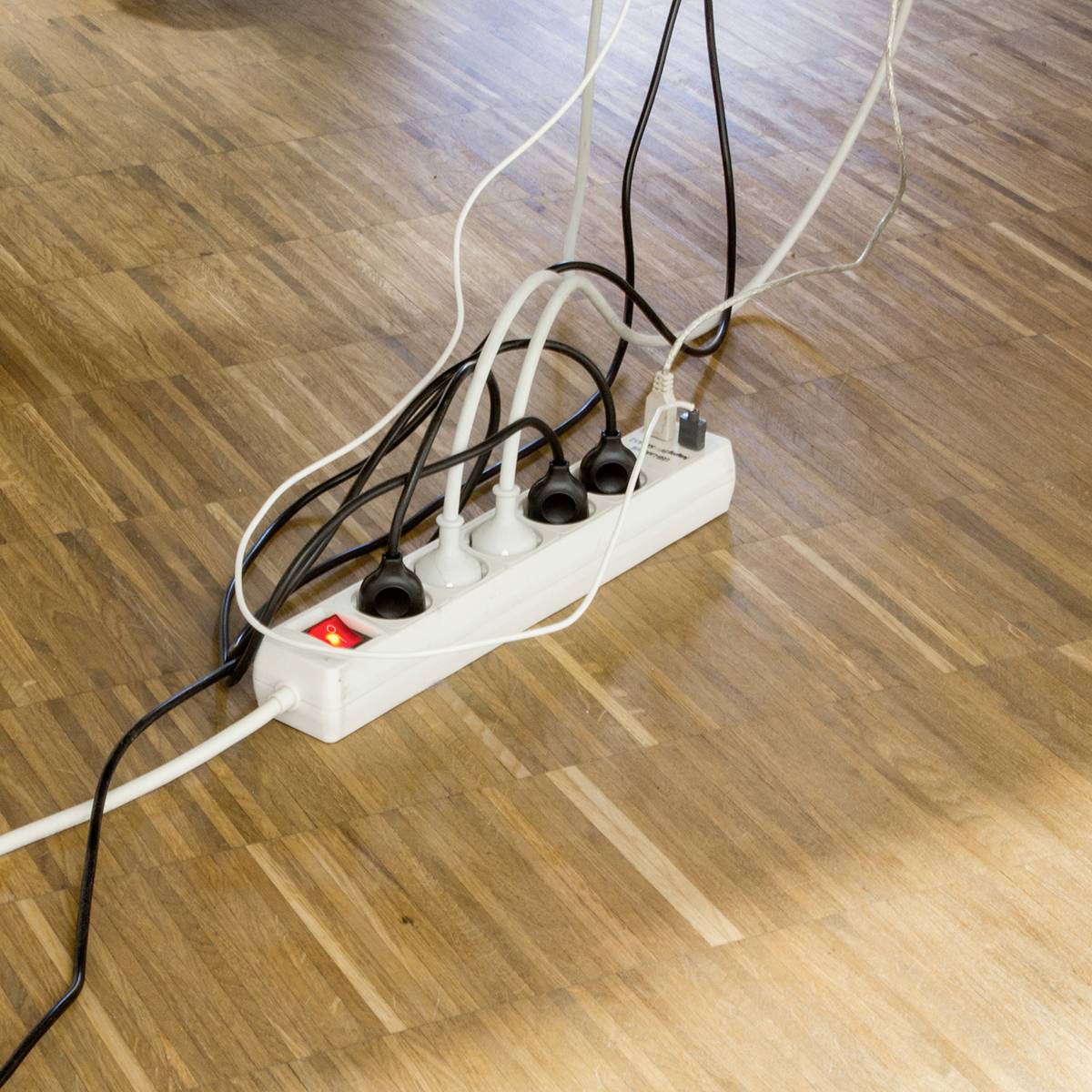 Mehrere elektrische Geräte sind an eine Steckdosenleiste auf einem Holzboden angeschlossen. Ein Schalter zeigt rot, was auf 
