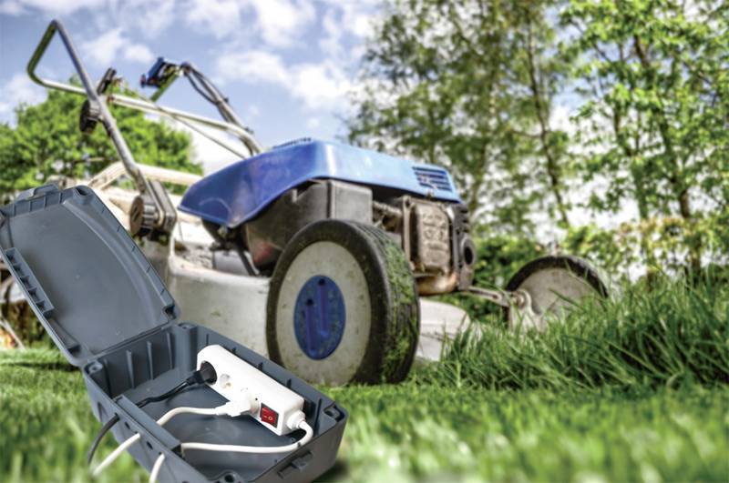 Ein blauer Rasenmäher steht auf hohem Gras im Garten, im Vordergrund liegt eine geöffnete Schutzhülle mit einem weißen Verlängerungskabel.