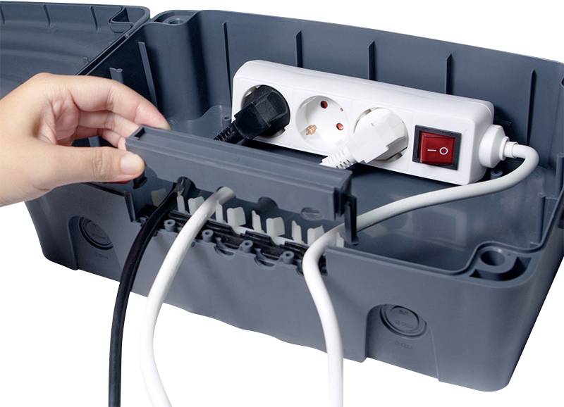 Eine Hand öffnet eine graue Plastikbox mit einem weißen Verlängerungskabel und mehreren eingesteckten schwarzen und weißen Kabeln.
