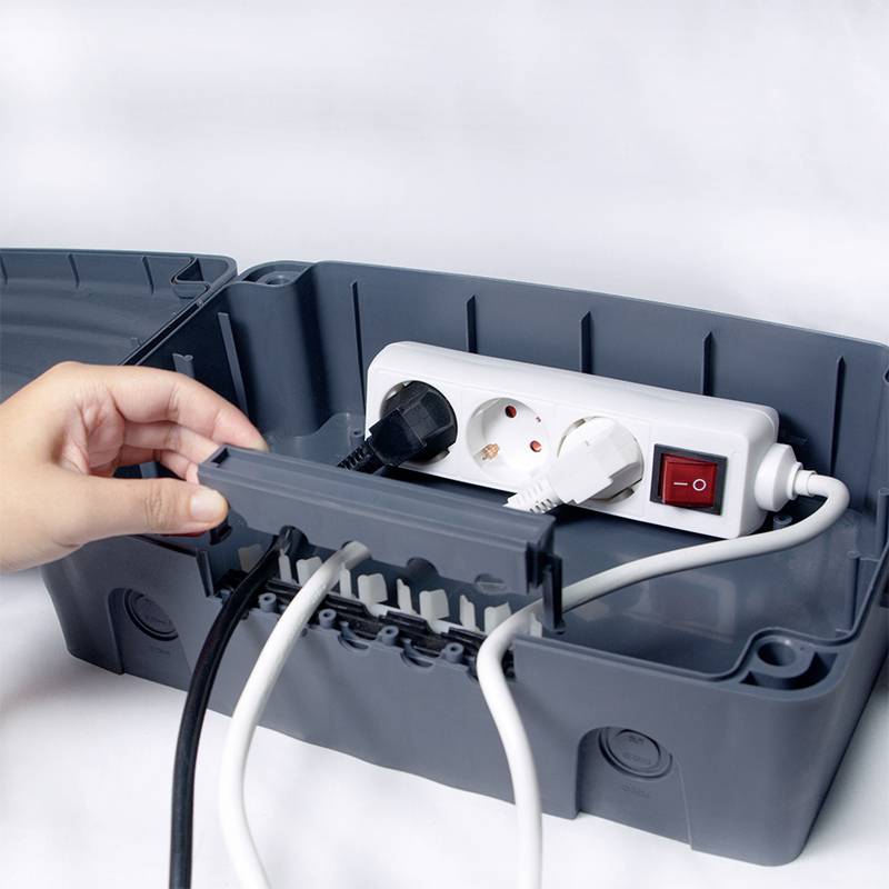 Eine Hand öffnet eine Kabelbox, in der sich eine Mehrfachsteckdose mit eingesteckten Kabeln und einem Netzschalter befindet.