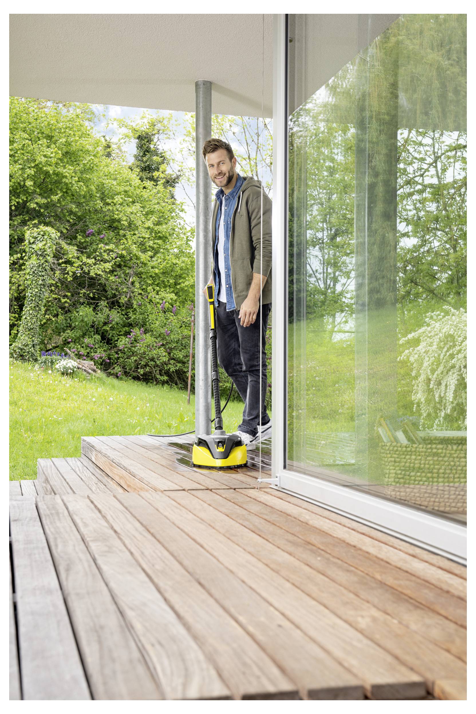 Eine Person verwendet einen gelben Hochdruckreiniger, um eine Holzterrasse außerhalb eines modernen Hauses zu reinigen, umgeben von Grünpflanzen und einer Glastür.