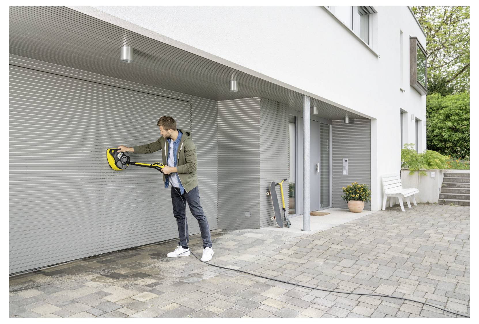Eine Person reinigt ein weißes Garagentor mit einem Hochdruckreiniger auf einer gepflasterten Einfahrt vor einem modernen Haus.