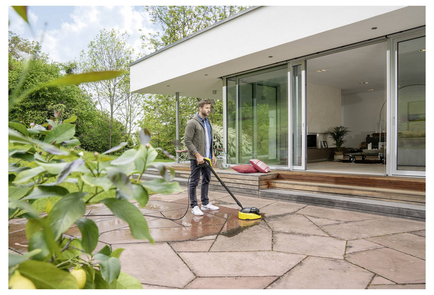 Eine Person reinigt eine große Steinveranda mit einem Hochdruckreiniger vor einem modernen Haus mit großen Glastüren. Büsche und Bäume umgeben das Areal.