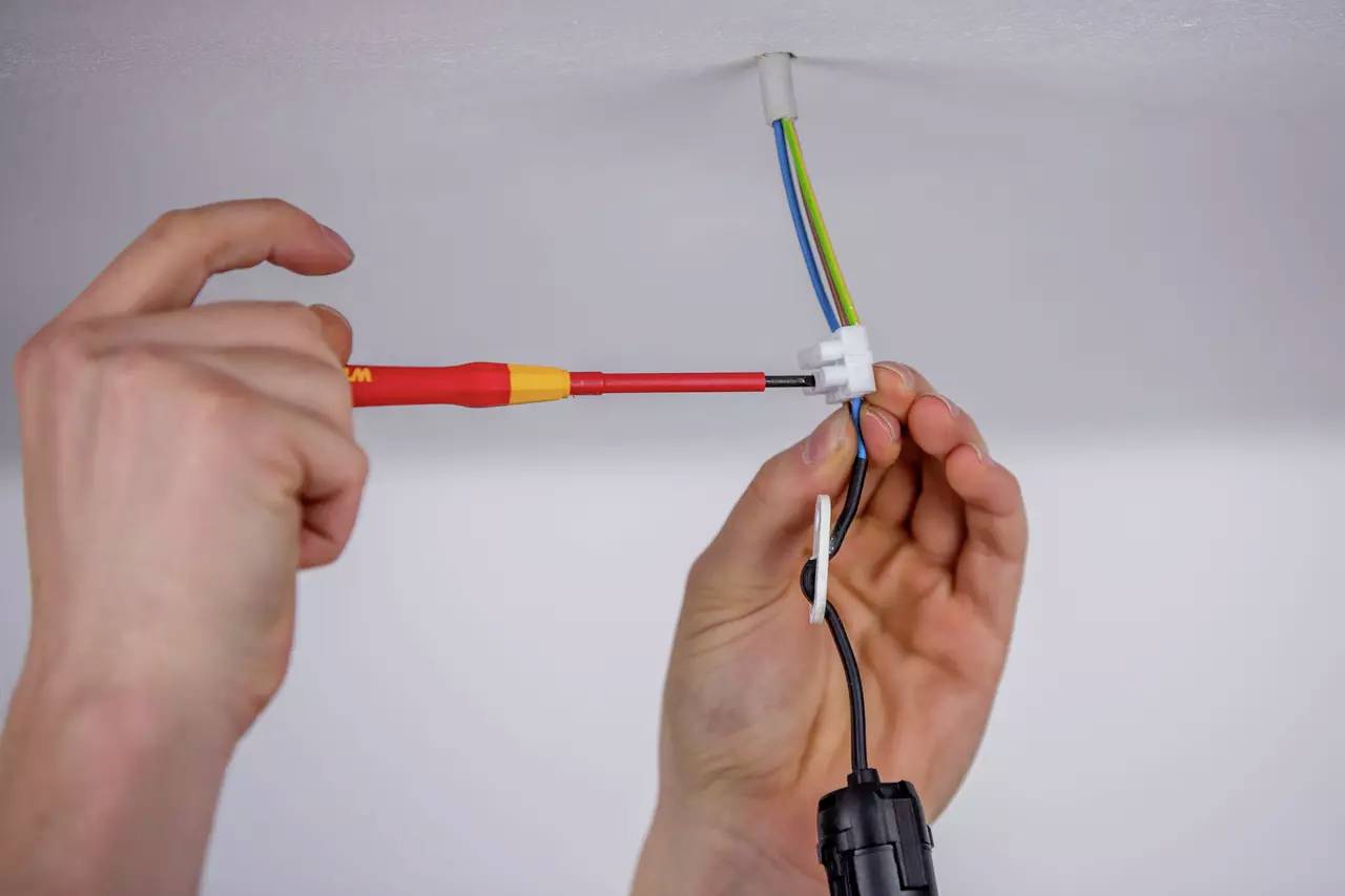 Person hält einen Schraubendreher und verbindet Kabel in einer elektrischen Anschlussdose an der Decke.
