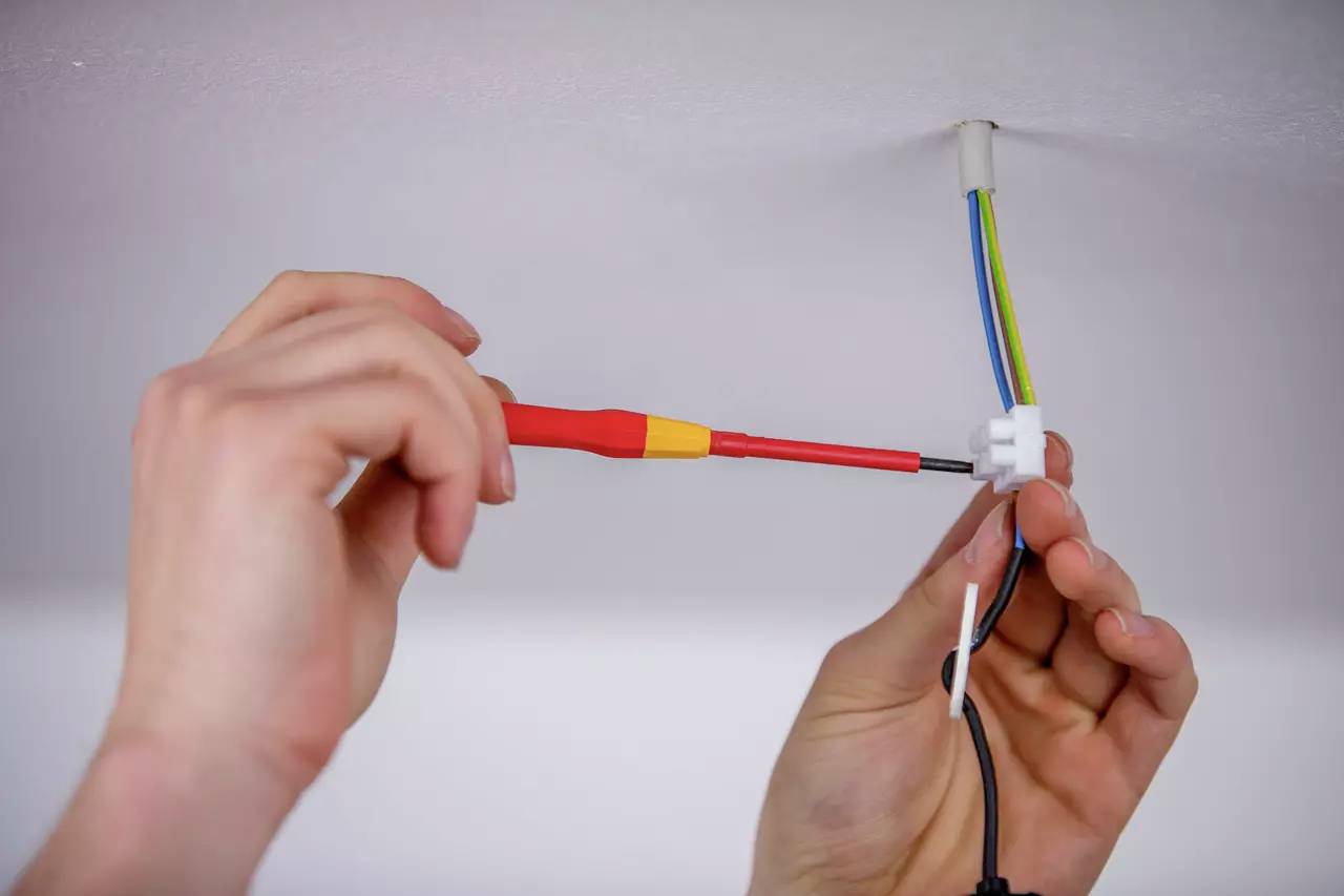 Einige Hände installieren ein elektrisches Kabel an der Decke. Ein Schraubendreher wird benutzt, um die Verbindungen zu sichern.