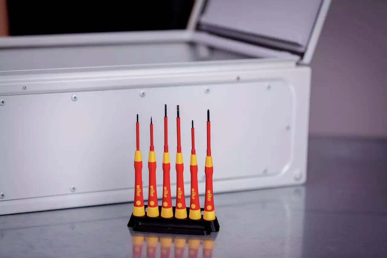 Sechs rote Schraubendreher in einem Ständer vor einem offenen, grauen Metallkasten auf einem Tisch.