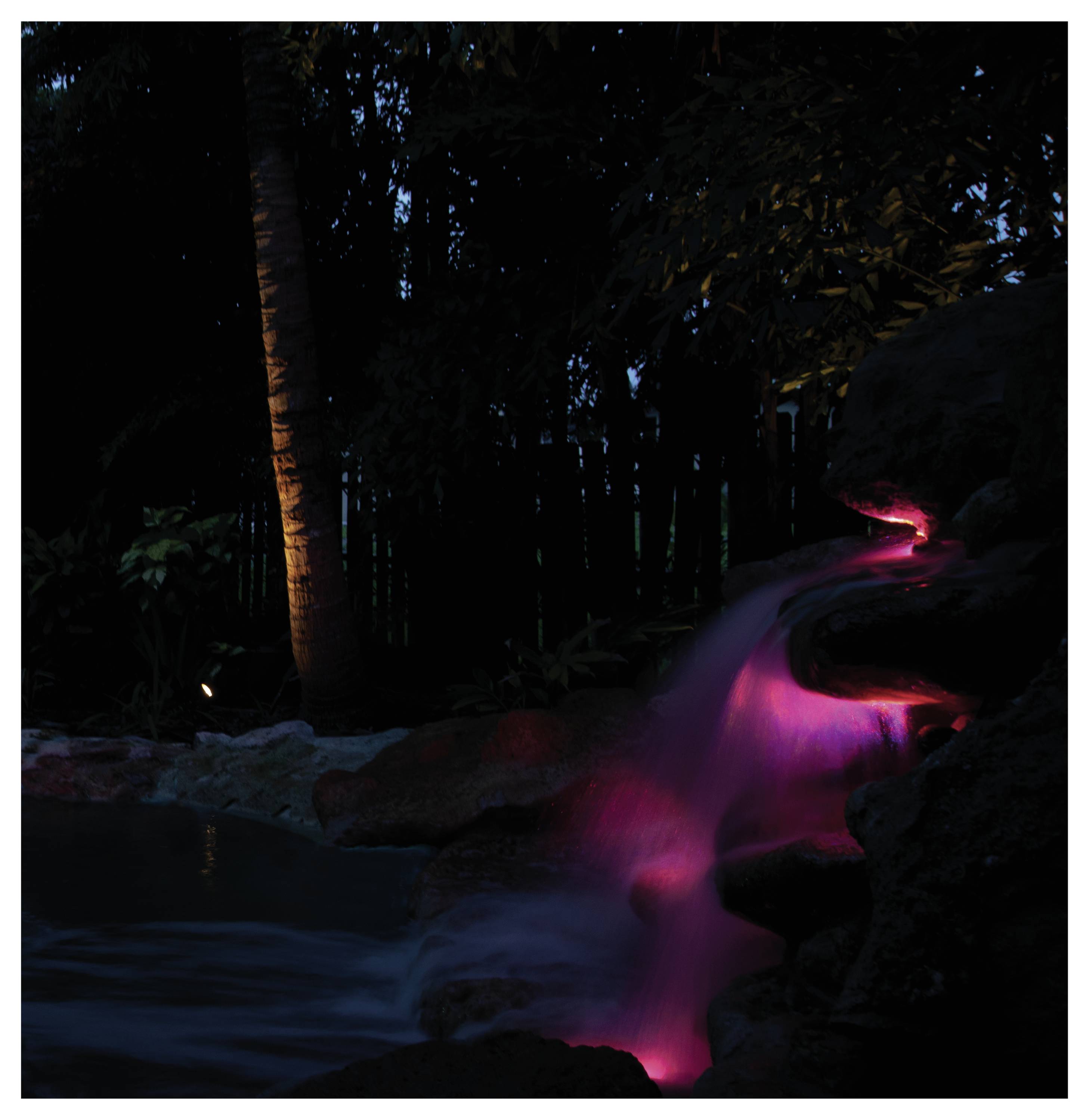 Wasserfall, beleuchtet von rosa Lichtern in einer dunklen Waldkulisse, der eine ruhige und mystische Atmosphäre erzeugt.