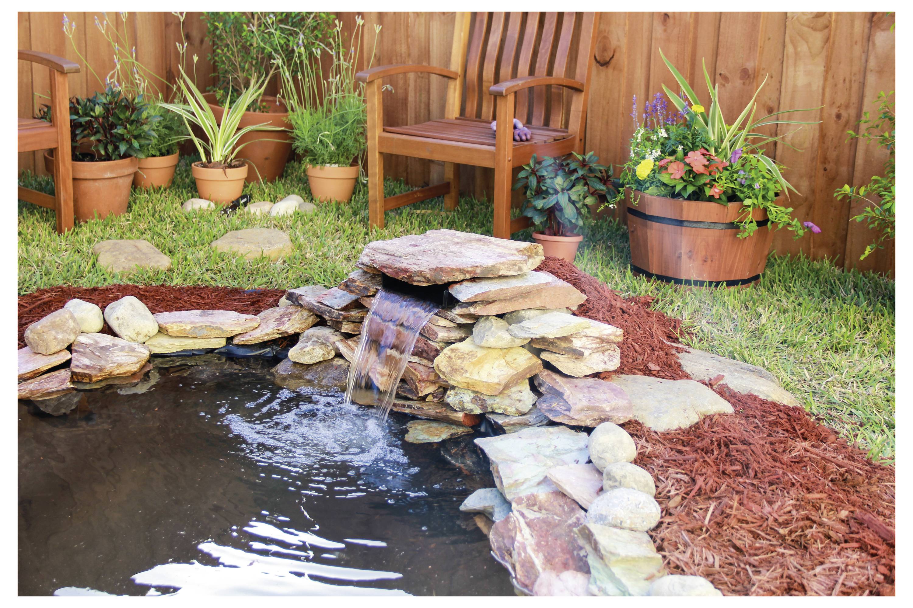 Ein kleiner Gartenteich mit einem Felsenwasserfall, umgeben von Mulch und Topfpflanzen. Holzstühle und ein Fassenpflanzer befinden sich in der Nähe.