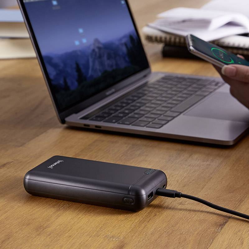 Laptop auf einem Holztisch; daneben eine Powerbank, die ein Smartphone lädt. Im Hintergrund Bücher.