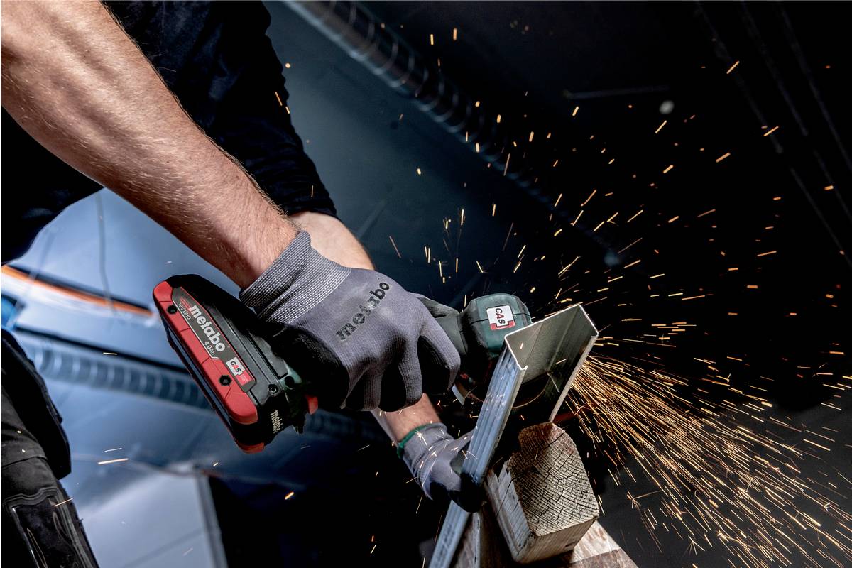 Eine Person schleift ein Metallstück mit einem Handwerkzeug, Funken sprühen, in einer Werkstattumgebung. Sie trägt Arbeitshandschuhe.