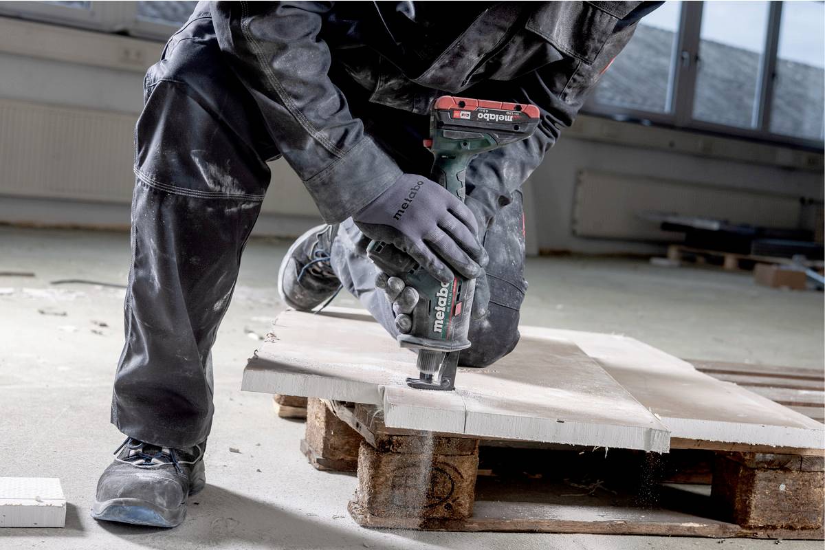Eine Person bearbeitet ein Holzbrett mit einem Elektrowerkzeug in einer Werkstatt. Die Person trägt Arbeitskleidung und Handschuhe.