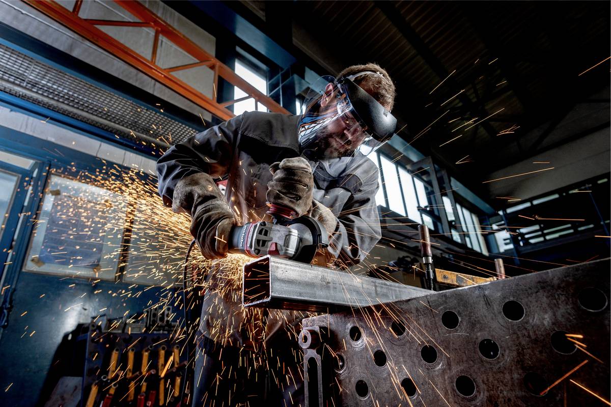 Ein Arbeiter mit Schutzbrille und Helm schleift ein Metallstück in einer Werkstatt. Funken fliegen von der Schleifmaschine.