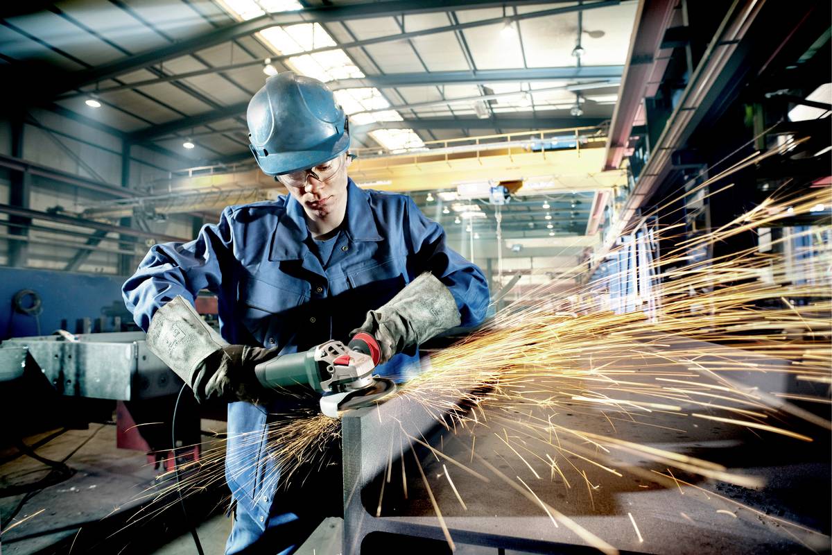 Ein Arbeiter in Schutzkleidung schleift Metall in einer Fabrik. Funken fliegen, während er konzentriert bei der Arbeit ist.