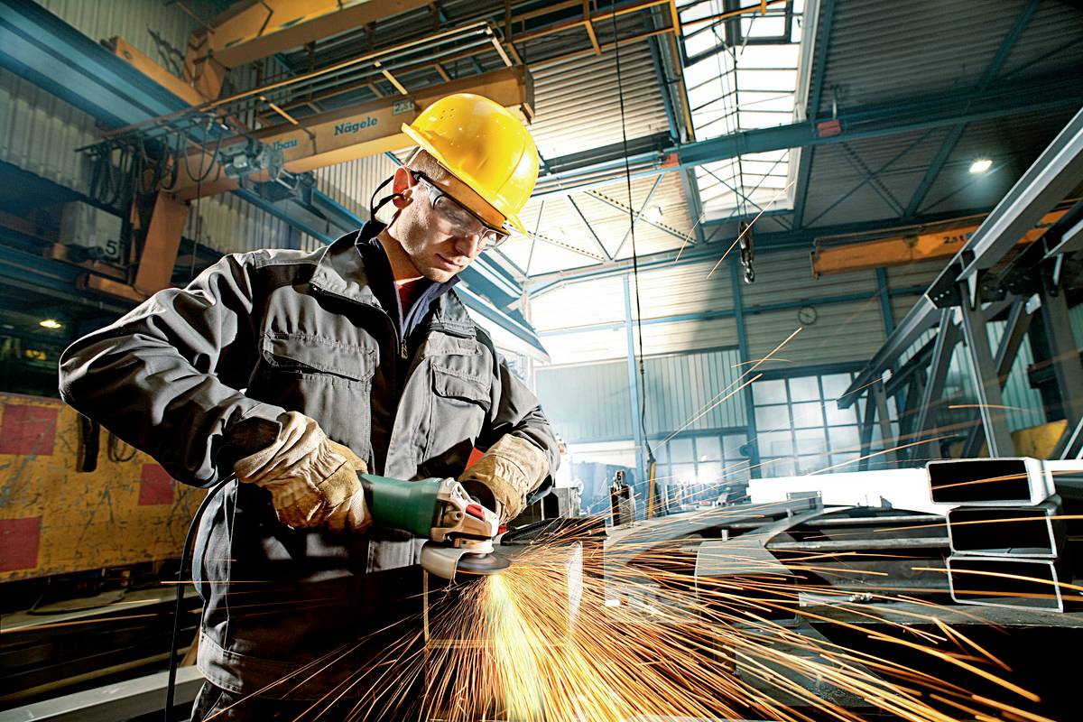 Ein Fabrikarbeiter mit Schutzhelm und -brille schleift mit einer Flexmaschine Metall, während Funken fliegen.