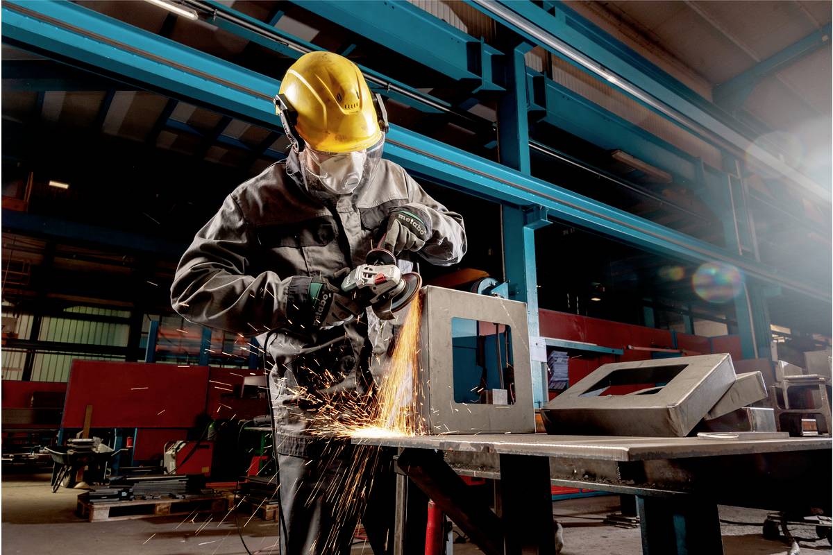 Ein Arbeiter in Schutzkleidung und Helm schleift Metall mit einem Winkelschleifer. Funken fliegen in einer industriellen Umgebung.