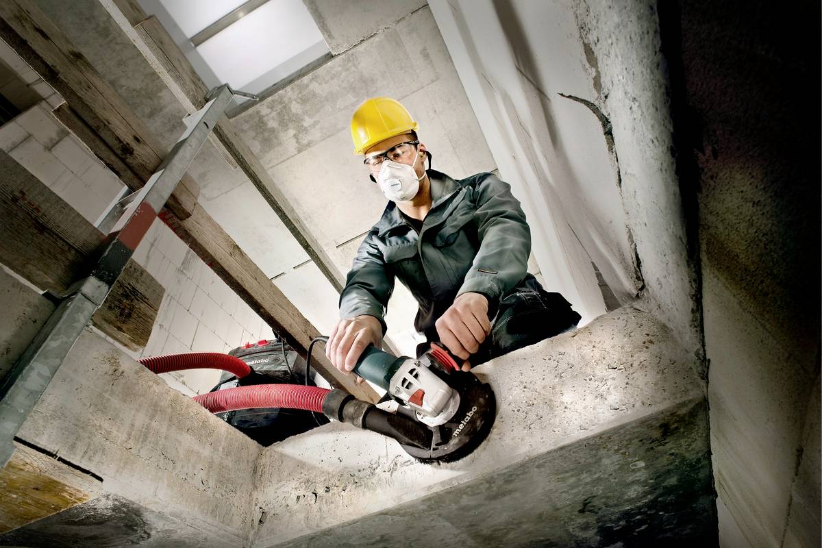 Arbeiter mit Schutzbrille, Helm und Staubmaske nutzt Winkelschleifer an Betonwand.