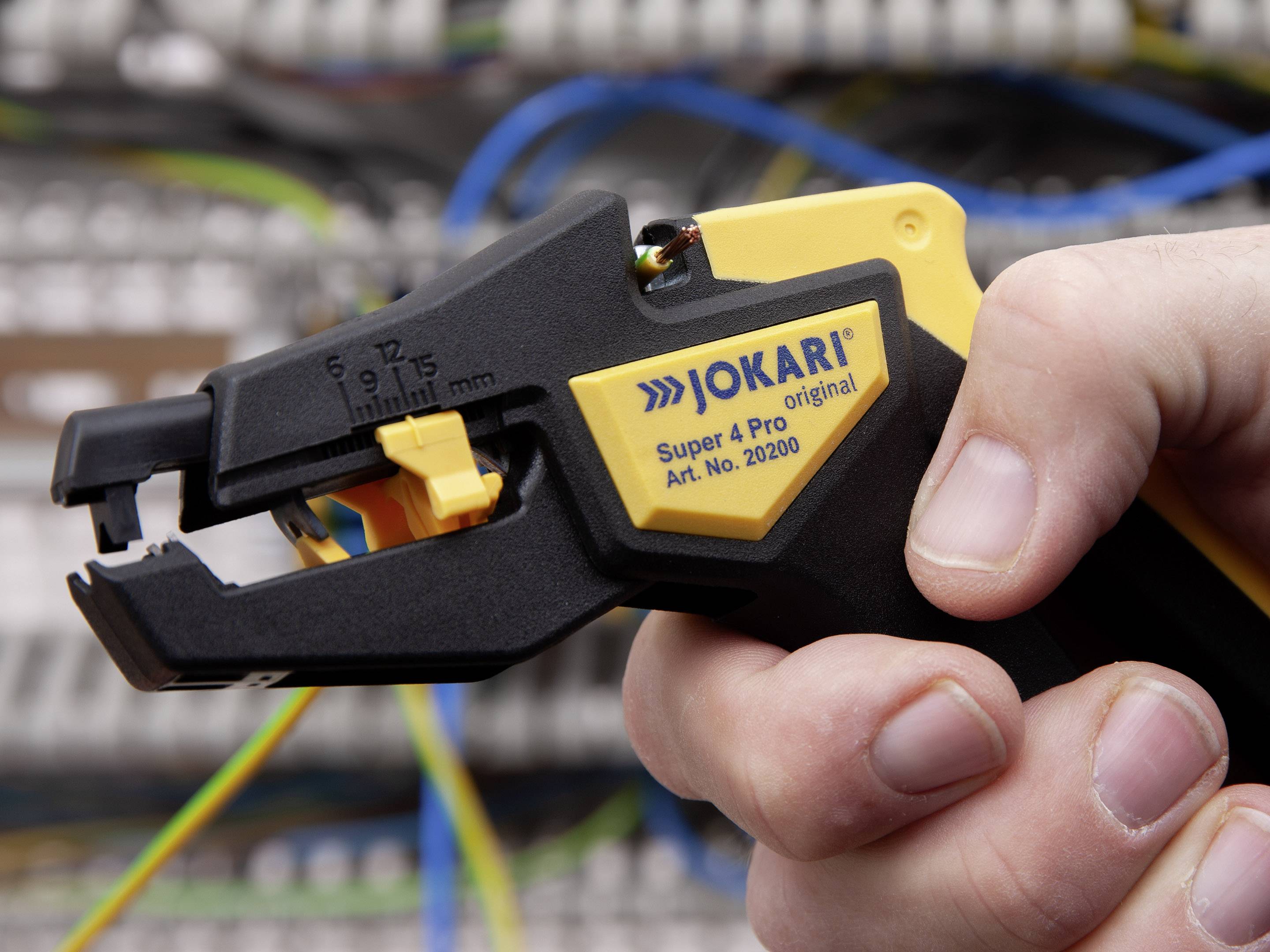 Hand hält einen Jokari Super 4 Pro Abisolierer vor einem Hintergrund mit elektrischen Kabeln.