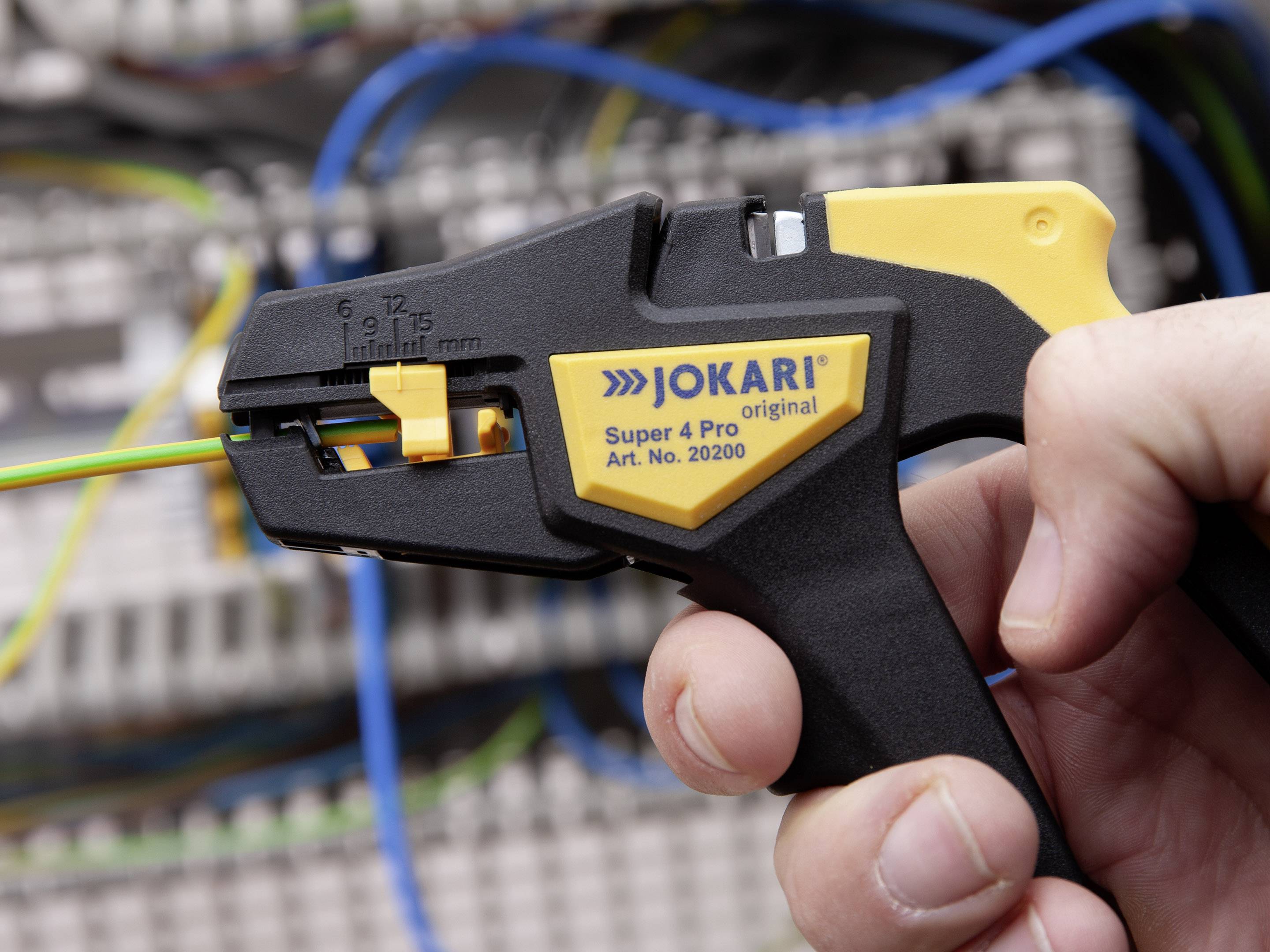 Eine Hand hält ein Abisolierwerkzeug vor einem unscharfen Hintergrund mit elektrischen Kabeln. Das Werkzeug ist für Kabelarbeiten geeignet.