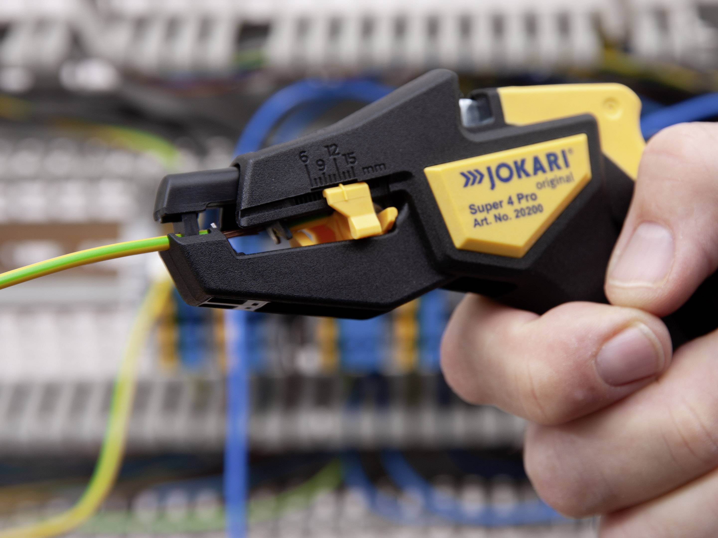 Eine Hand hält einen Abisolierer, um ein grün-gelbes Kabel abzuisolieren. Der Hintergrund zeigt unscharfe elektrische Schaltkästen.