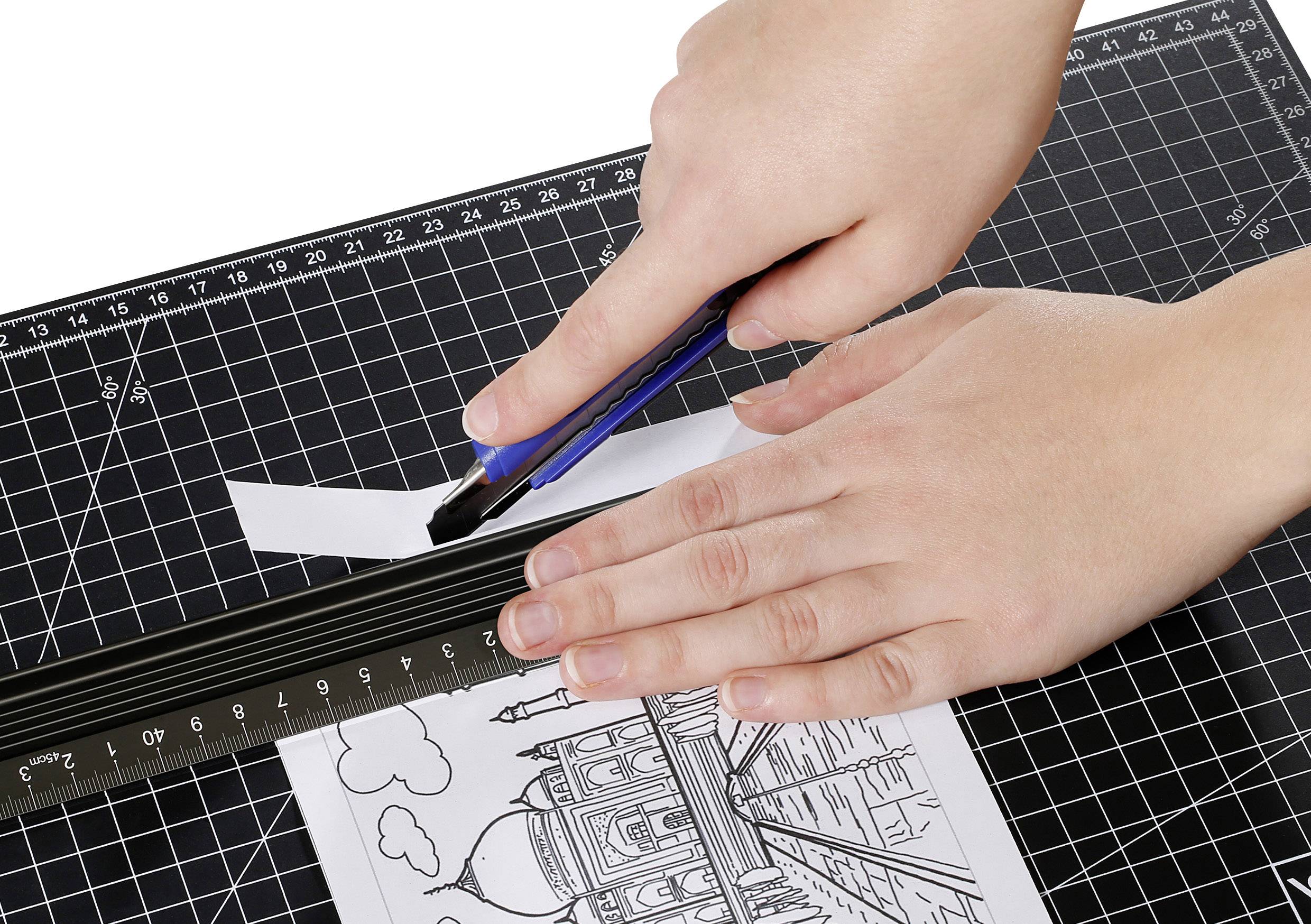 Hände schneiden Papier mit einem Cutter an einem Lineal entlang auf einer Schneidematte. Das Bild zeigt eine Architekturzeichnung.