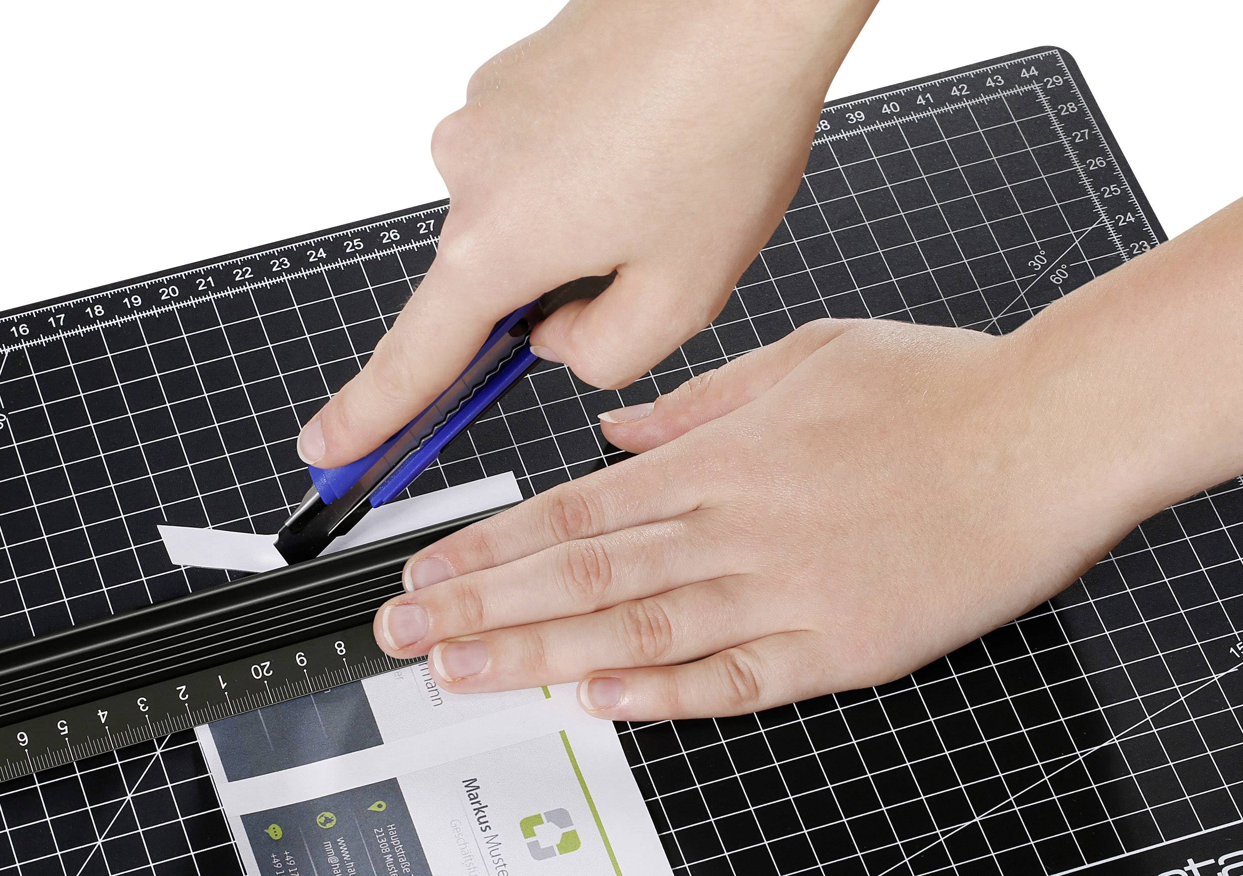 Zwei Hände schneiden mit einem Cuttermesser Papier auf einer schwarzen Schneidematte. Eine Hand hält ein Lineal, die andere führt das Messer.