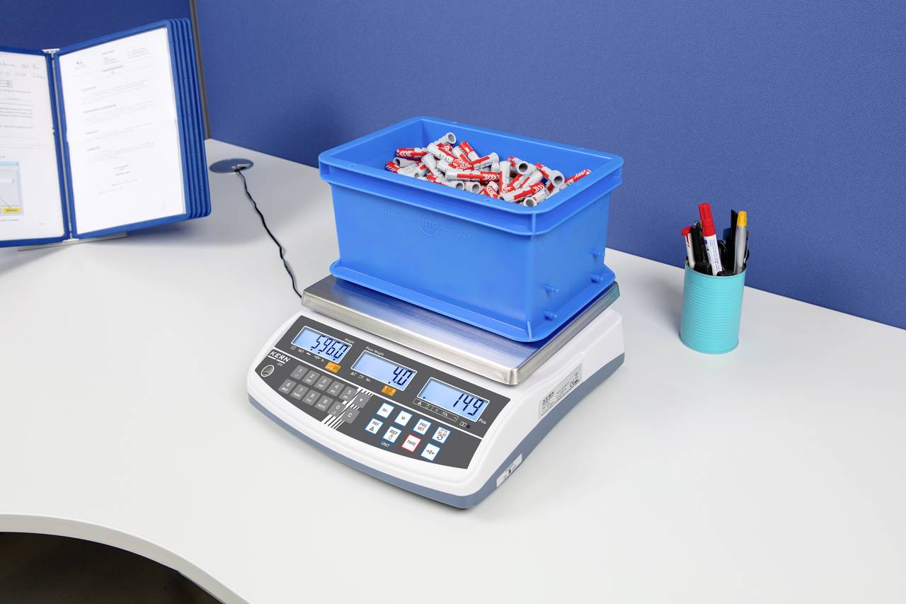 Blaue Box mit roten und weißen Teststreifen auf einer digitalen Waage, die 5,567 Kilogramm anzeigt. Im Hintergrund Büroartikel auf einem Tisch.