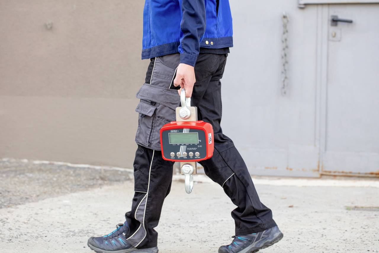 Eine Person in Arbeitshose und -jacke trägt ein rotes Messgerät mit Digitalanzeige. Im Hintergrund eine graue Tür.