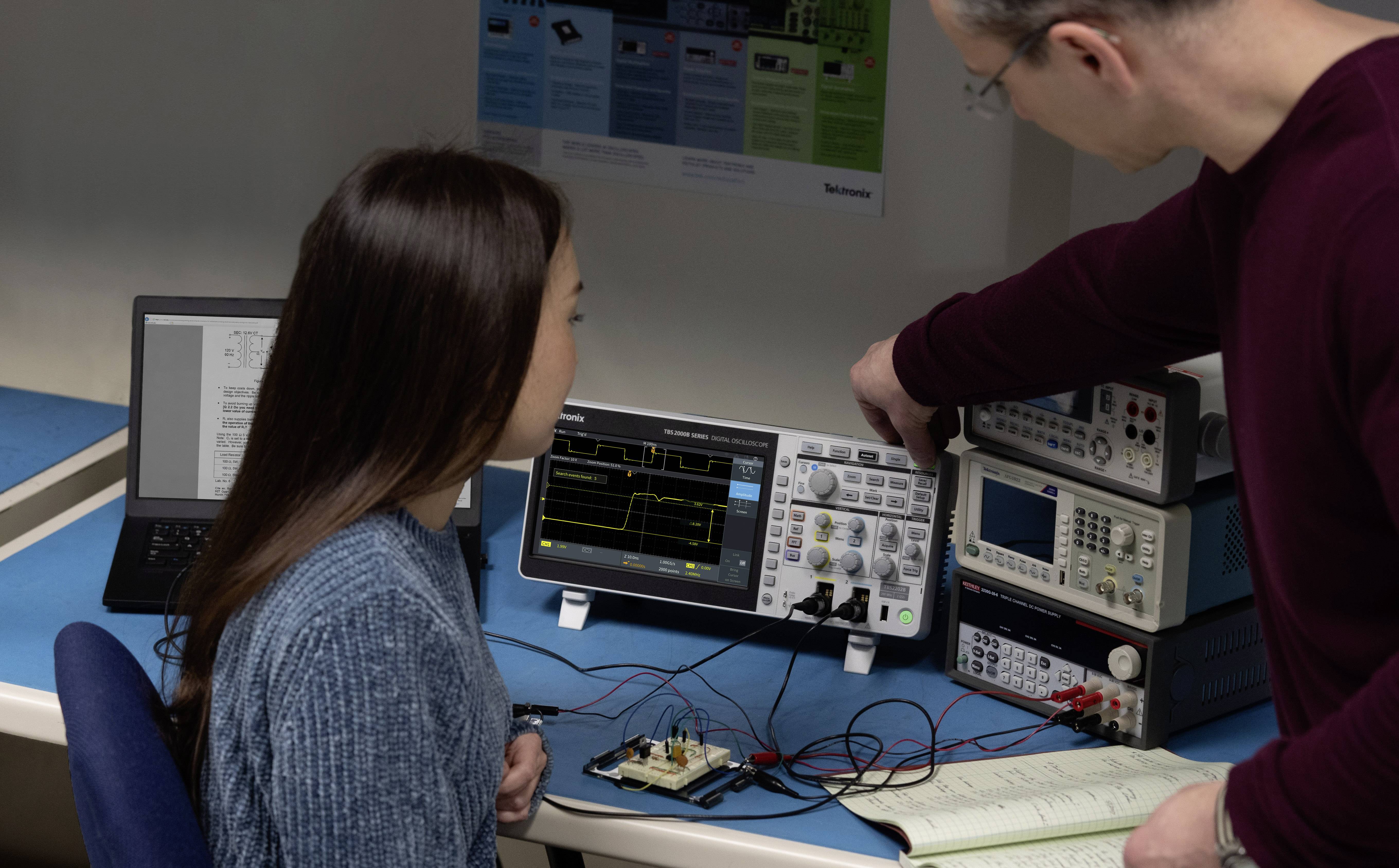 Ein Mann erklärt einer Frau die Verwendung eines Oszilloskops. Im Hintergrund sind Computer und elektronische Geräte sichtbar.