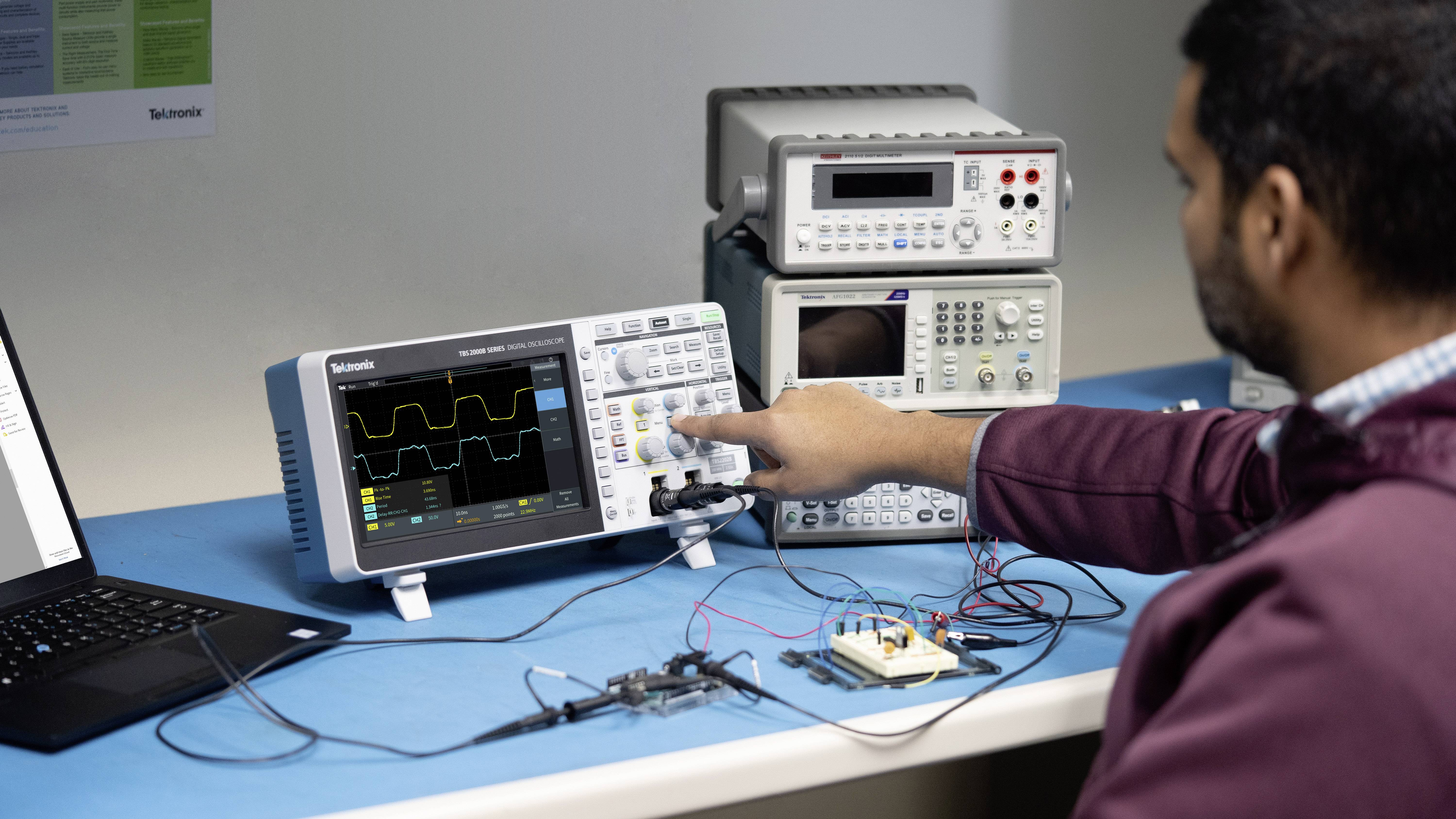 Ein Mann bedient ein Oszilloskop zur Messung elektrischer Signale an einem Schreibtisch, auf dem auch ein Laptop und andere Messgeräte stehen.