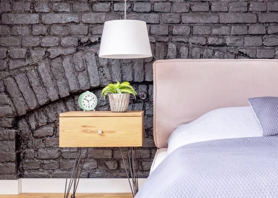 Nachtisch mit Wecker, Pflanze und Lampe neben einem Bett vor einer Backsteinwand.