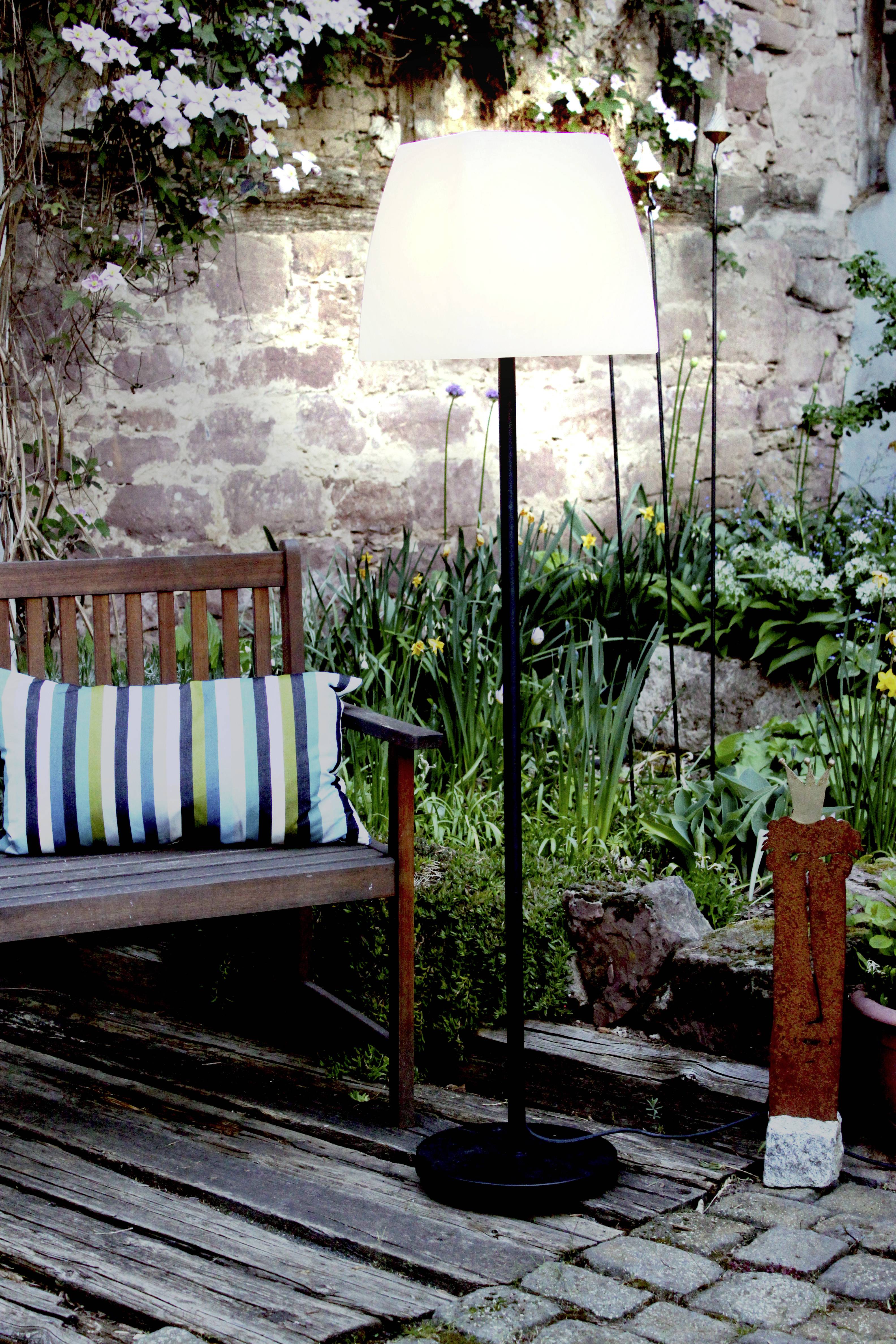 Stehlampe beleuchtet einen Gartensitzbereich mit Bank und Kissen, umgeben von Blumen und Pflanzen vor einer alten Steinmauer.