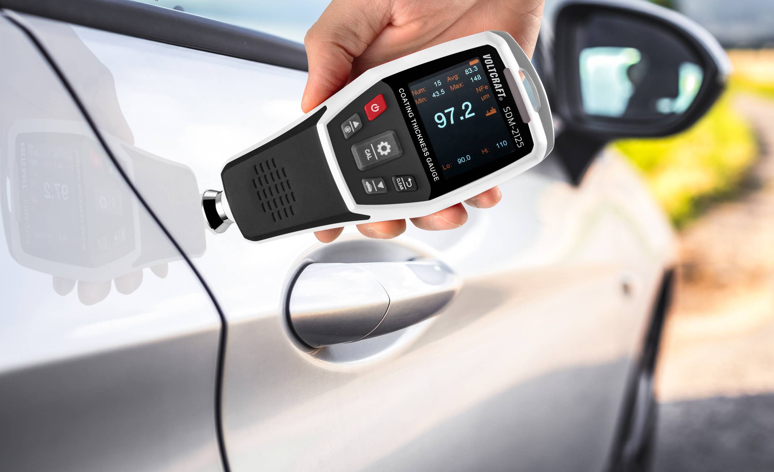 Eine Person hält ein Schichtdickenmessgerät an die Autotür, das Display zeigt '97.2' an. Das Auto ist hellgrau, Hintergrund ist unscharf.