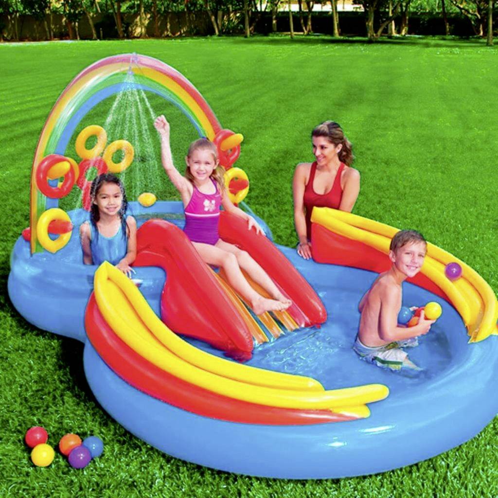Drei Kinder und eine Erwachsene spielen in einem aufblasbaren Planschbecken auf einer Wiese. Das Becken hat eine Rutsche und einen Regenbogen.