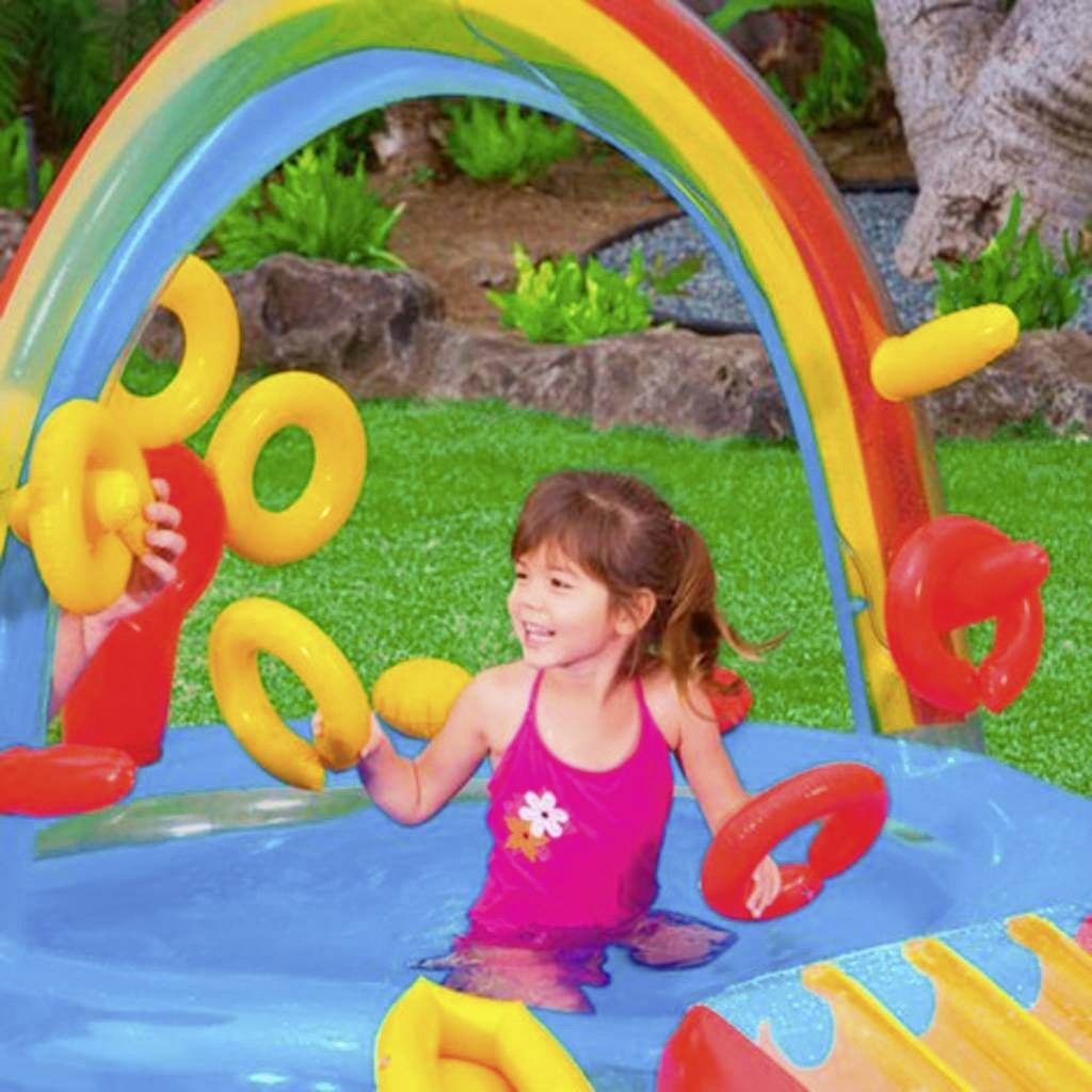 Ein Kind spielt fröhlich in einem bunten Wasserspielplatz mit aufblasbaren Ringen und einem Regenbogen-Dekor im Hintergrund.