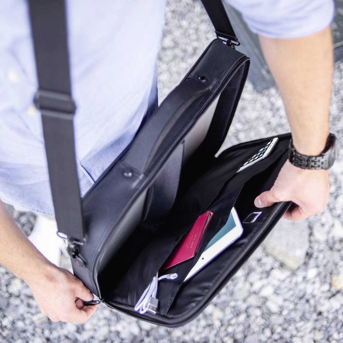 Person öffnet schwarze Umhängetasche mit Laptop, Smartphone und Notizbuch darin, auf einem Kiesweg.