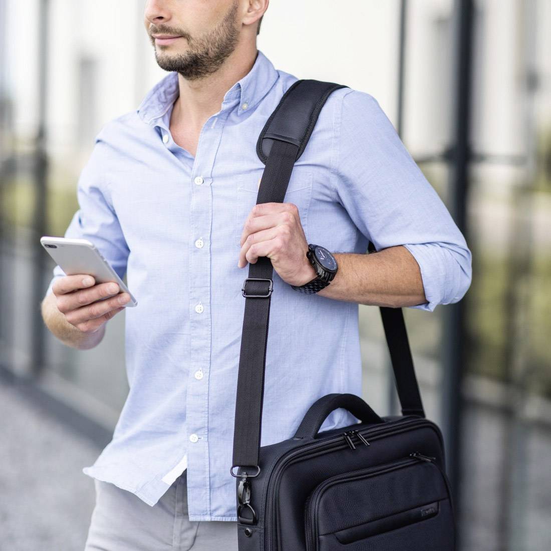 Ein Mann mit Bart und geschlossenem, hellblauem Hemd steht draußen, hält ein Smartphone und trägt eine Umhängetasche.