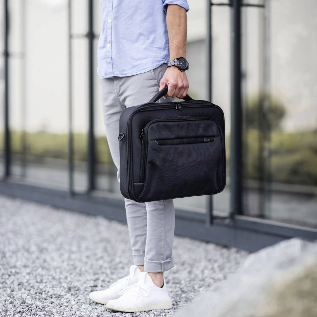 Eine Person trägt eine schwarze Umhängetasche und steht auf Schotter vor einem modernen, verglasten Gebäude.