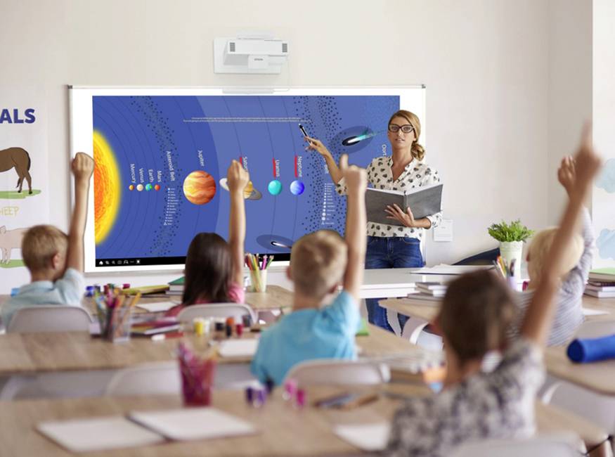 Eine Lehrerin erklärt mit einem digitalen Whiteboard das Sonnensystem in einer Klasse. Schüler sitzen und heben die Hand.