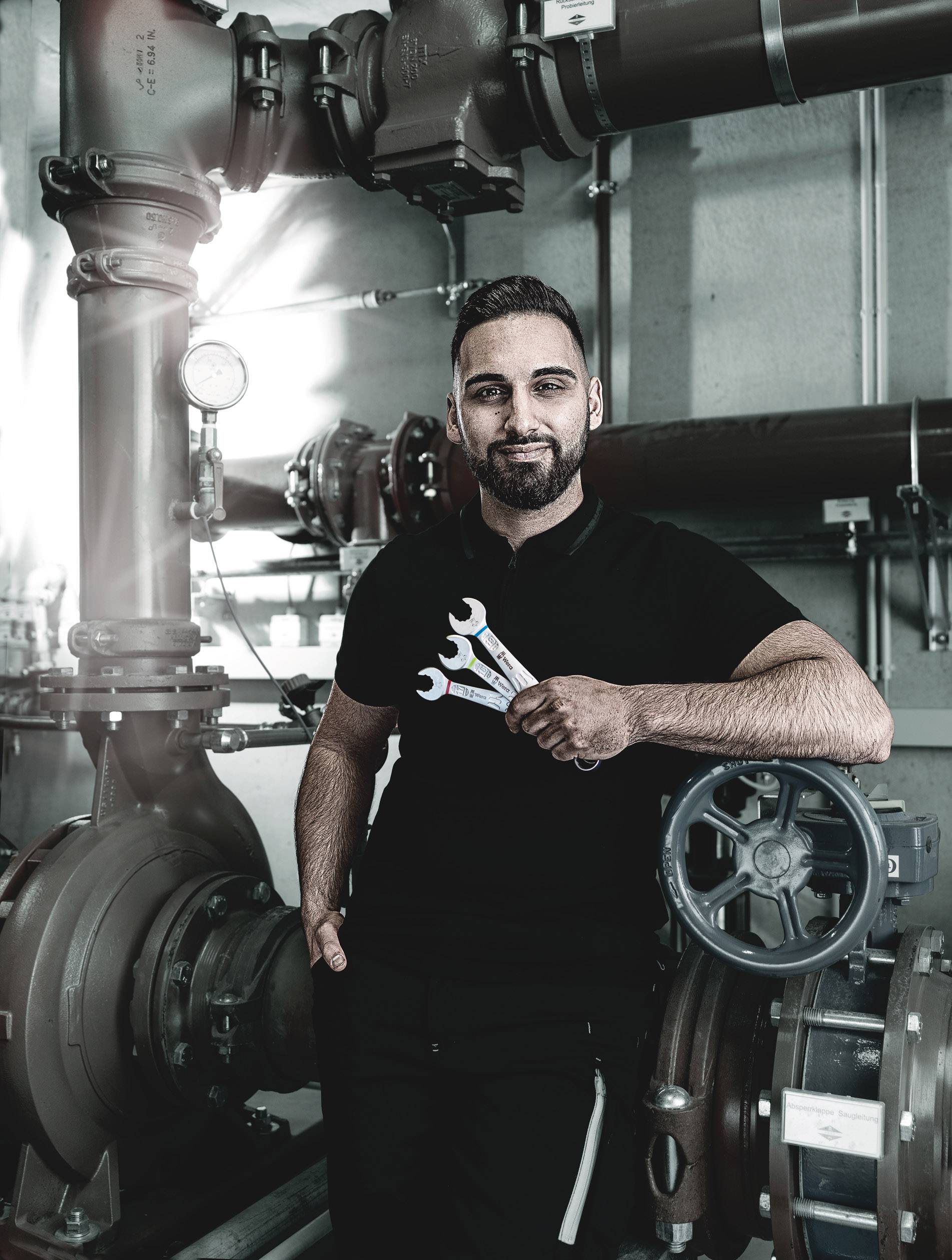 Eine Person in Arbeitskleidung steht in einem Industrieraum mit Rohren und Ventilen, hält drei Schraubenschlüssel in der Hand.