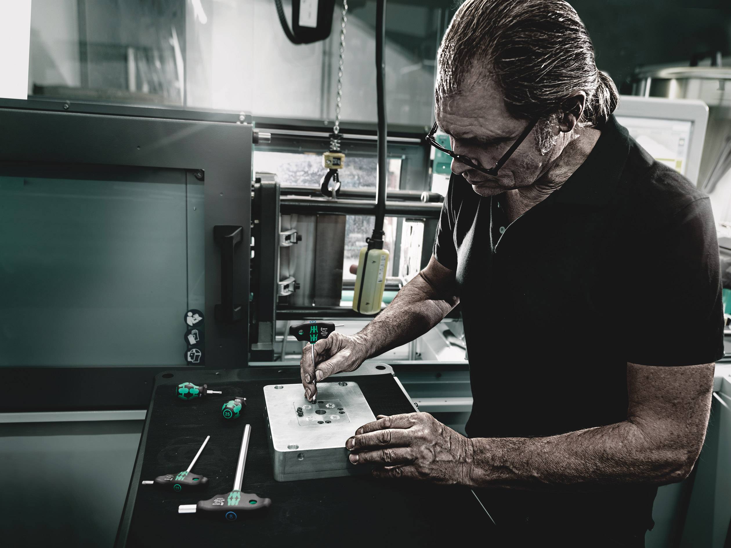 Ein Mann arbeitet in einer Werkstatt, er bearbeitet sorgfältig ein Metallteil auf einem Tisch mit verschiedenen Werkzeugen um ihn herum.