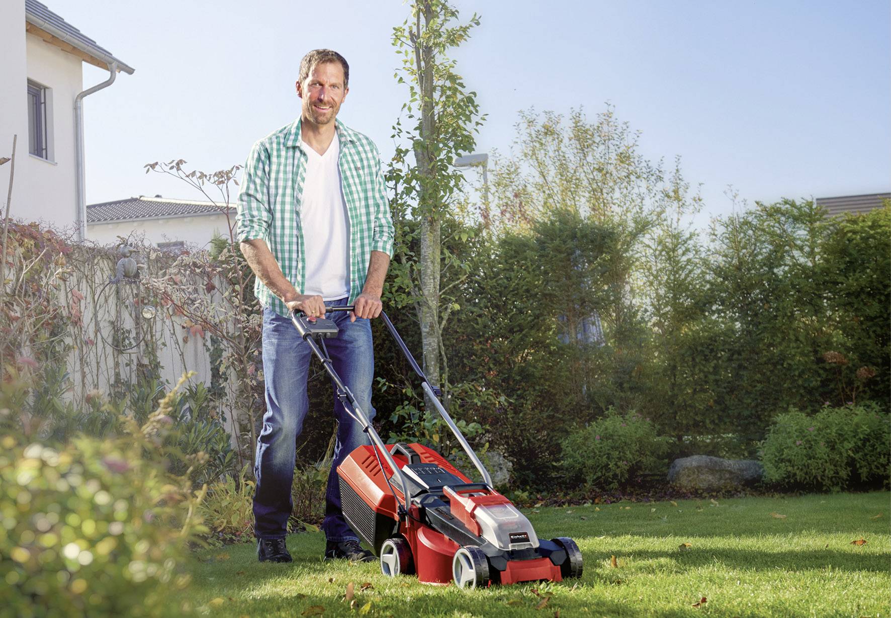 Ein Mann mäht lächelnd den Rasen mit einem roten Rasenmäher an einem sonnigen Tag in einem grünen Garten.