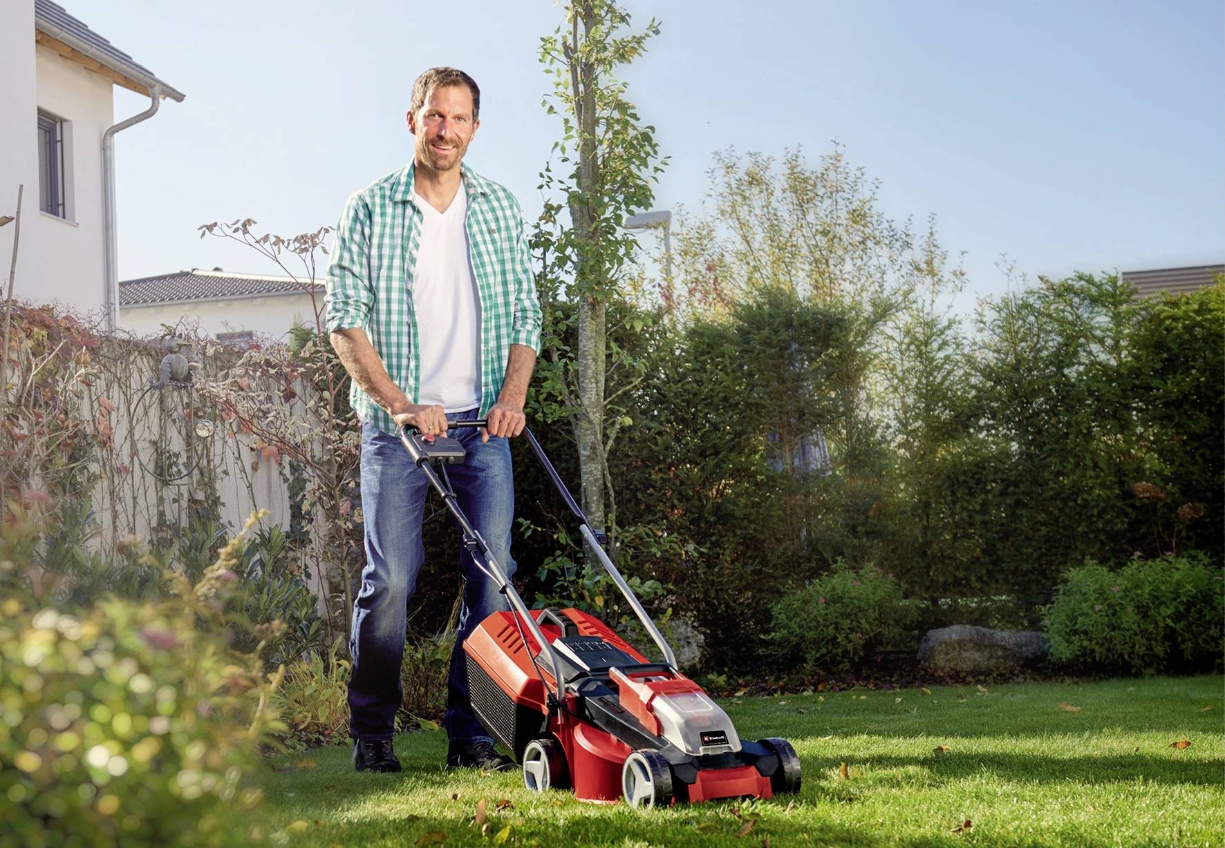 Ein Mann mäht bei sonnigem Wetter den Rasen im Garten mit einem roten Rasenmäher, umgeben von Büschen und einem Haus im Hintergrund.