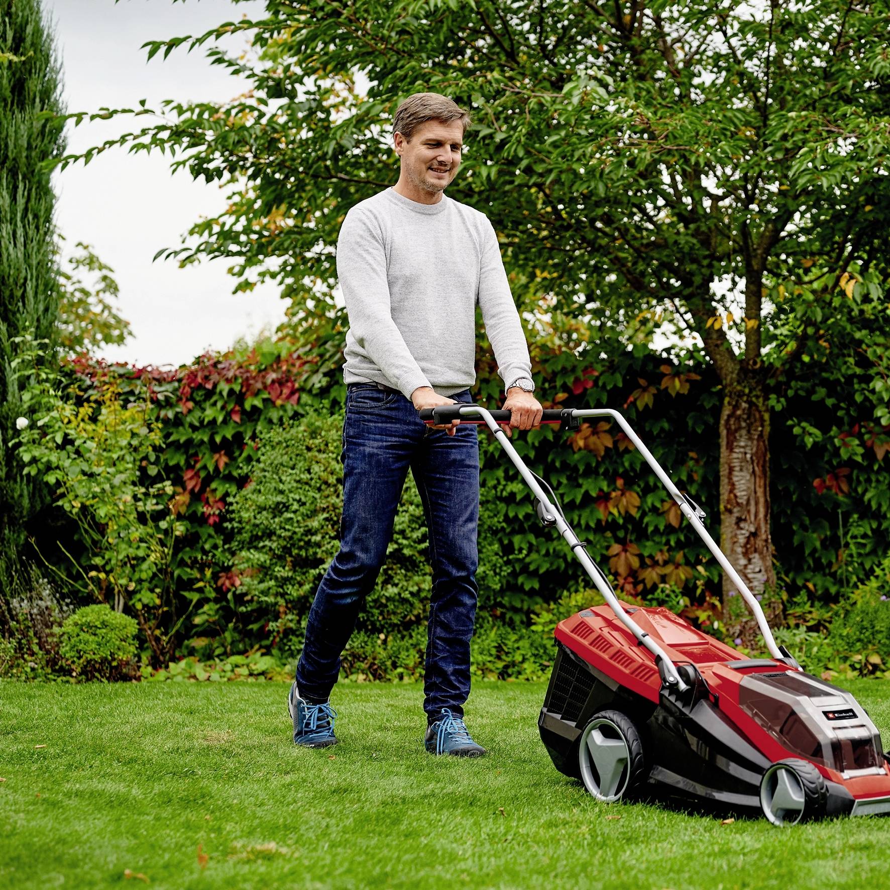 Ein Mann mäht den Rasen in einem Garten. Er trägt einen grauen Pullover und blaue Schuhe. Im Hintergrund sind Bäume und Sträucher zu sehen.