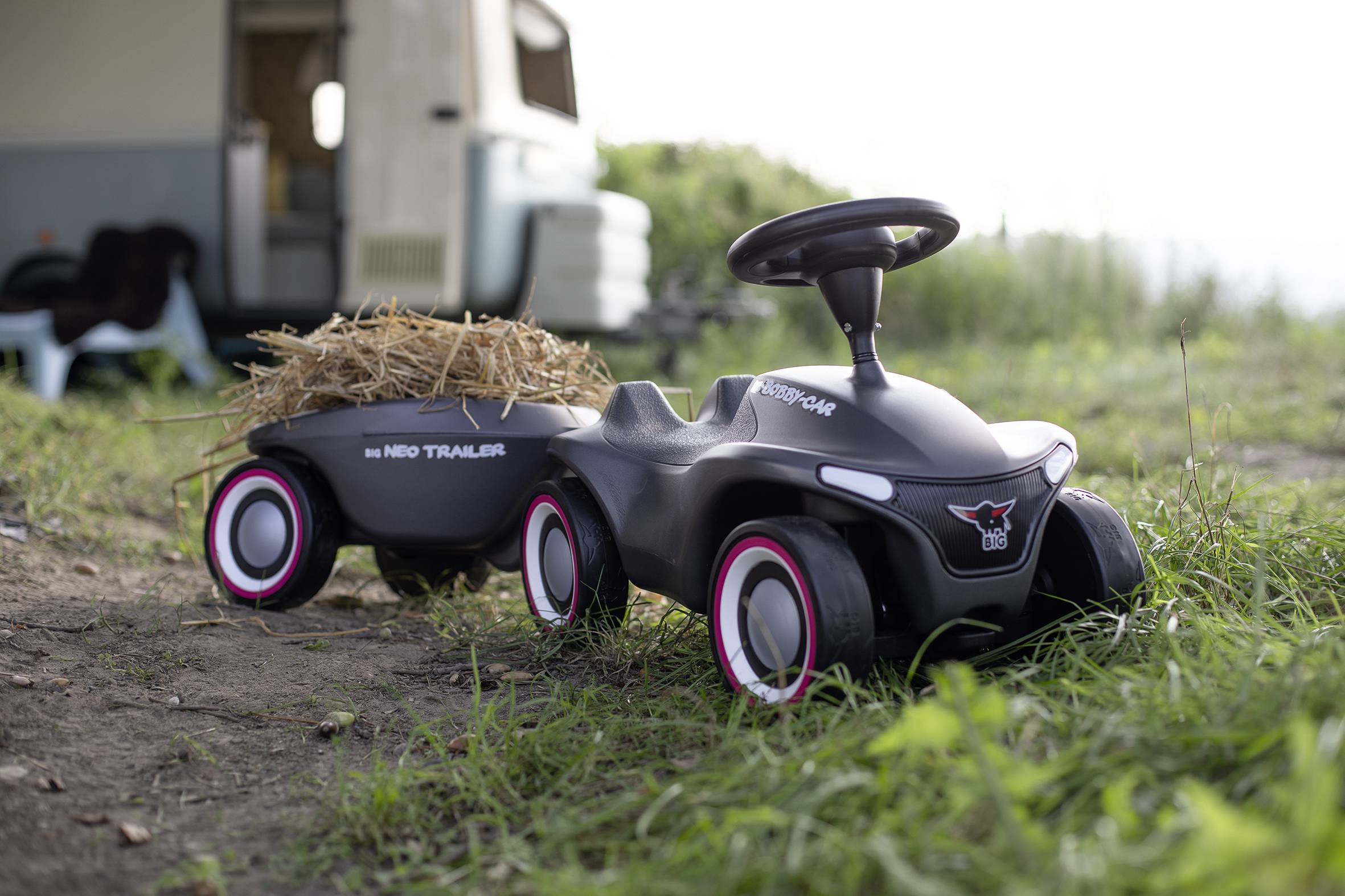 Spielzeugauto mit Anhänger auf einer Wiese, beladen mit Heu. Im Hintergrund ein geparkter Wohnwagen.